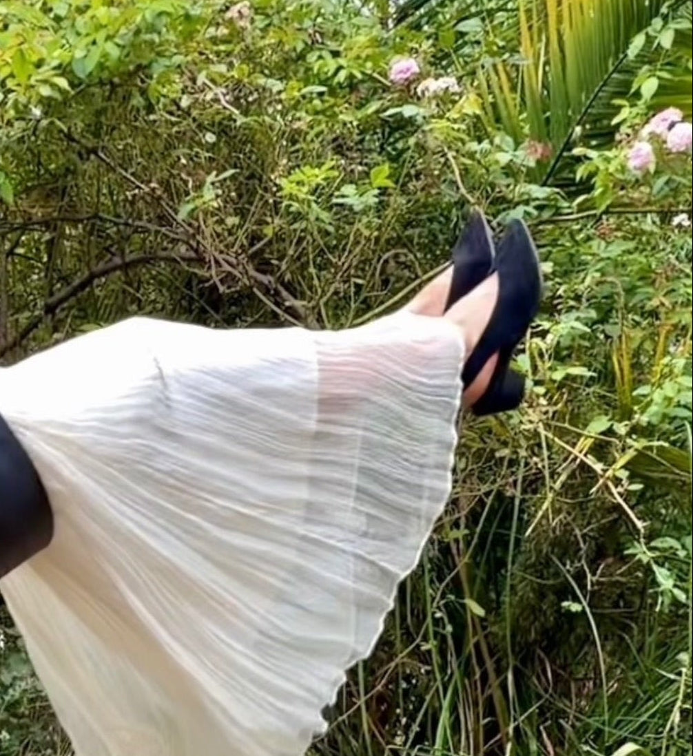 A person outdoors in a lush green garden, wearing a long pleated white skirt, black high heels, and a wide black belt over a brown and black patterned top. A swing set is visible on the left, and tropical plants fill the background.
